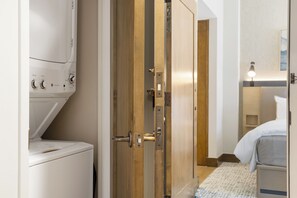 Une chambre d’hôtel équipée d’un lave-linge, d’une porte en bois, d’un lit et d’une table de chevet avec une lampe.