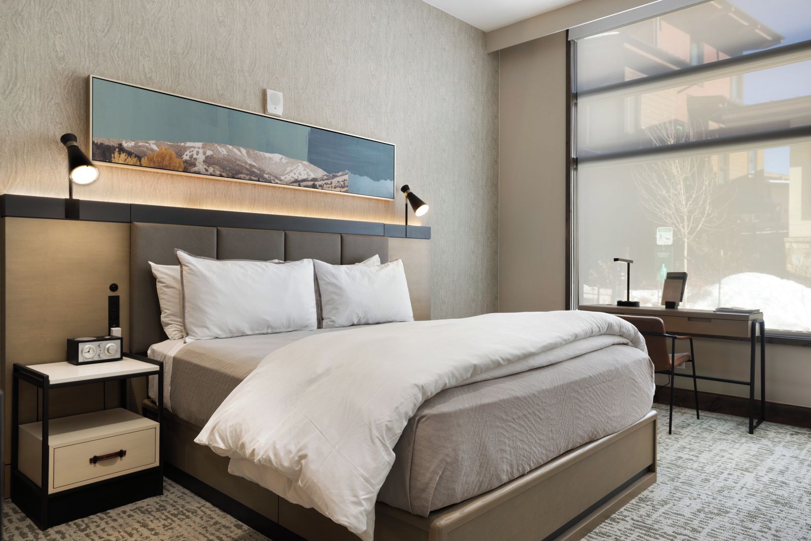 A modern hotel room with a large bed, bedside tables, a desk, and a large window with a view of the city.