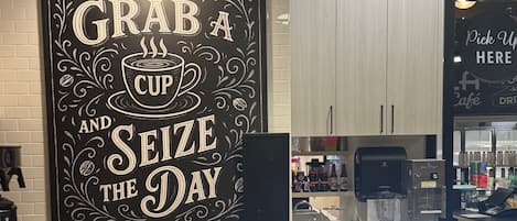 A coffee shop interior with a wall sign that reads "GRAB A CUP AND SEIZE THE DAY," a coffee machine, a display case with various drinks, and a counter with a menu.