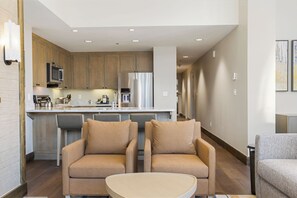 Une cuisine moderne avec des meubles en bois, des appareils en acier inoxydable et un îlot central. Le salon comprend deux fauteuils beiges et une petite table ronde.