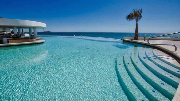 A clear blue infinity pool with a palm tree and a modern building structure in the background. - Las Palmas 2BR 2BA Suite 2nd FL Ocean View G205 (Puerto Peñasco)