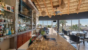 A bar area with a thatched roof, a large TV screen, and a view of the ocean through large windows. - Las Palmas 2BR 2BA Suite 2nd FL Ocean View G205 (Puerto Peñasco)