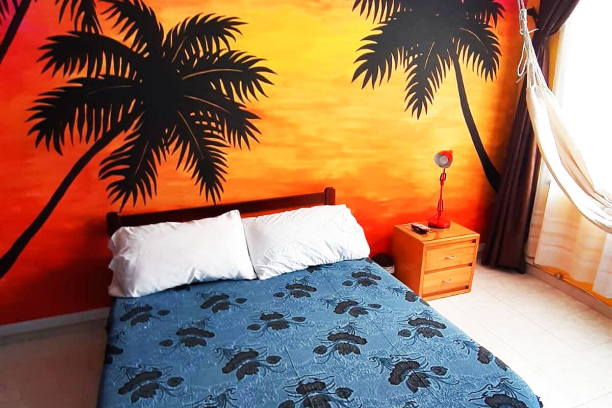Ein Bett mit blauer Blumenbettdecke, einem Holz-Kopfschild und weißen Kissen. Ein Nachttisch mit Lampe und ein kleiner Tisch befinden sich neben dem Bett. An der Wand ist eine farbenfrohe Wandmalerei mit Palmen zu sehen.