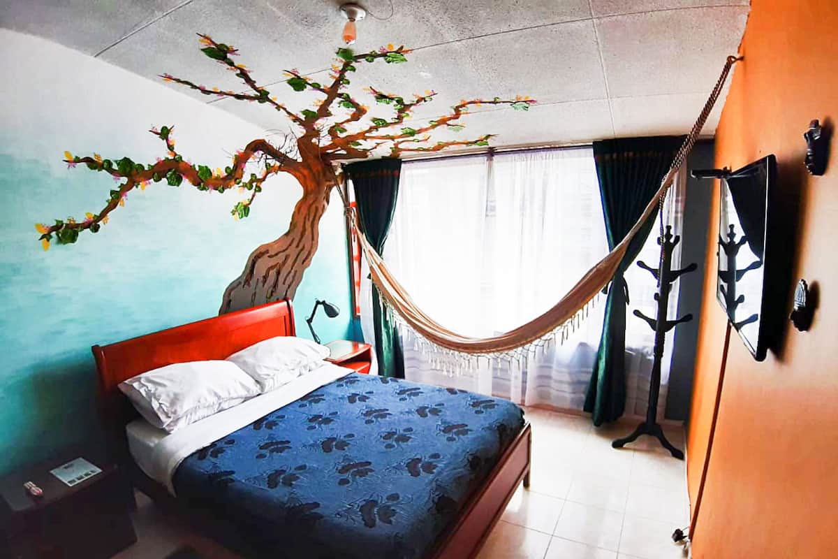 Ein Schlafzimmer mit Bett, einer Hängematte, einer Baum-Wandmalerei, einem Fenster mit Vorhängen und einem Kleiderständer.