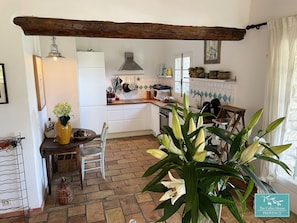 Una cocina con armarios blancos, una viga de techo de madera, un colgador para ollas y una ventana con cortinas.