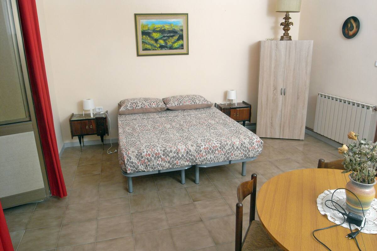 A bedroom with a bed, bedside tables, a wardrobe, a dining table with chairs, and a vase with flowers.