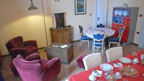 Un coin repas avec une nappe rouge, des assiettes blanches et une radio ancienne. On y trouve des fauteuils violets et un buffet en bois rempli de vaisselle.