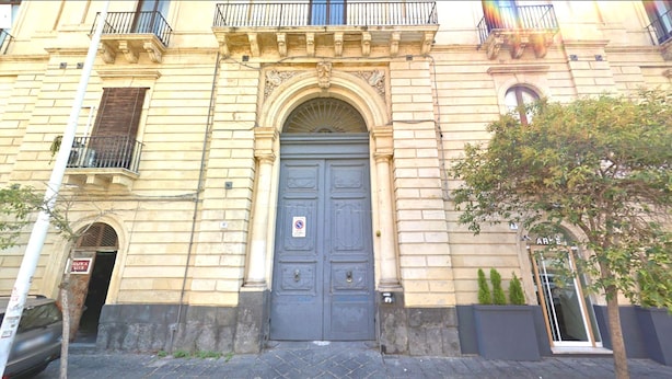 Un bâtiment doté d’une grande porte bleue au centre, d’un balcon avec une rambarde en métal et d’un petit arbre planté dans un pot.