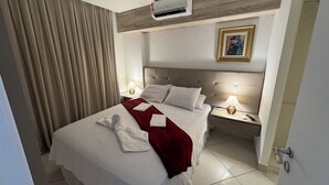 A hotel room with a large bed, two bedside tables with lamps, an air conditioning unit above the bed, a painting on the wall, and a door to the right. - Landscape Beira Mar, Lindo, Luxo, Vista mar (Fortaleza)