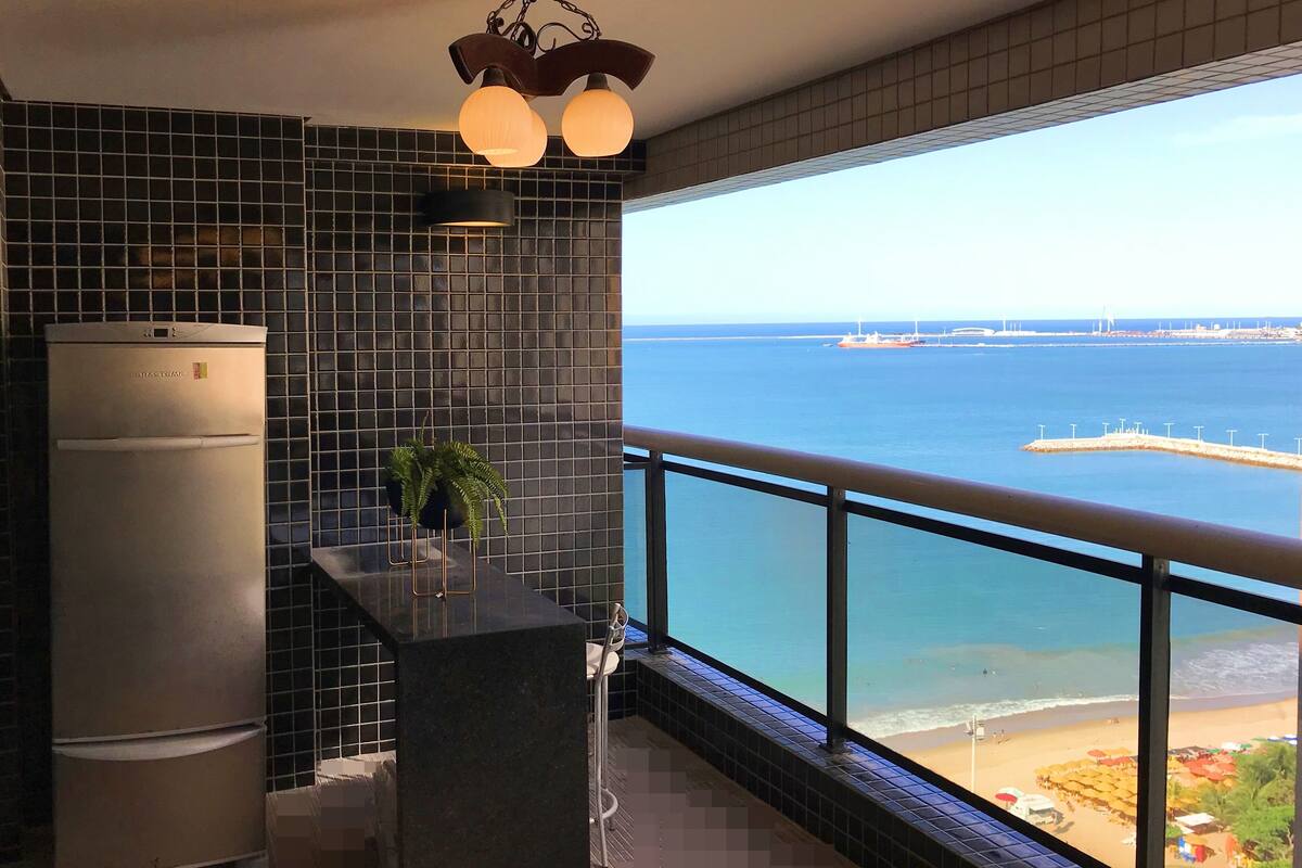 A kitchen with a view of the sea, a refrigerator, a potted plant, and a bar area.