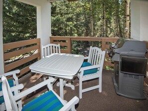 Een overdekt terras met een witte tafel en stoelen, een barbecue en uitzicht op een bebost gebied.