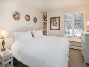 Een keurig opgemaakte slaapkamer met een bed, nachtkastjes, een boekenkast en een raam met jaloezieën.