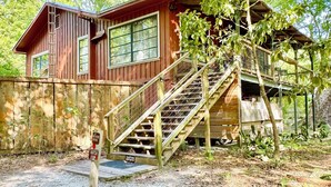 A rustic wooden house with a staircase leading to a balcony, surrounded by trees and a gravel ground. - Bowman's Landing Spring House on the Santa Fe River (Fort White)