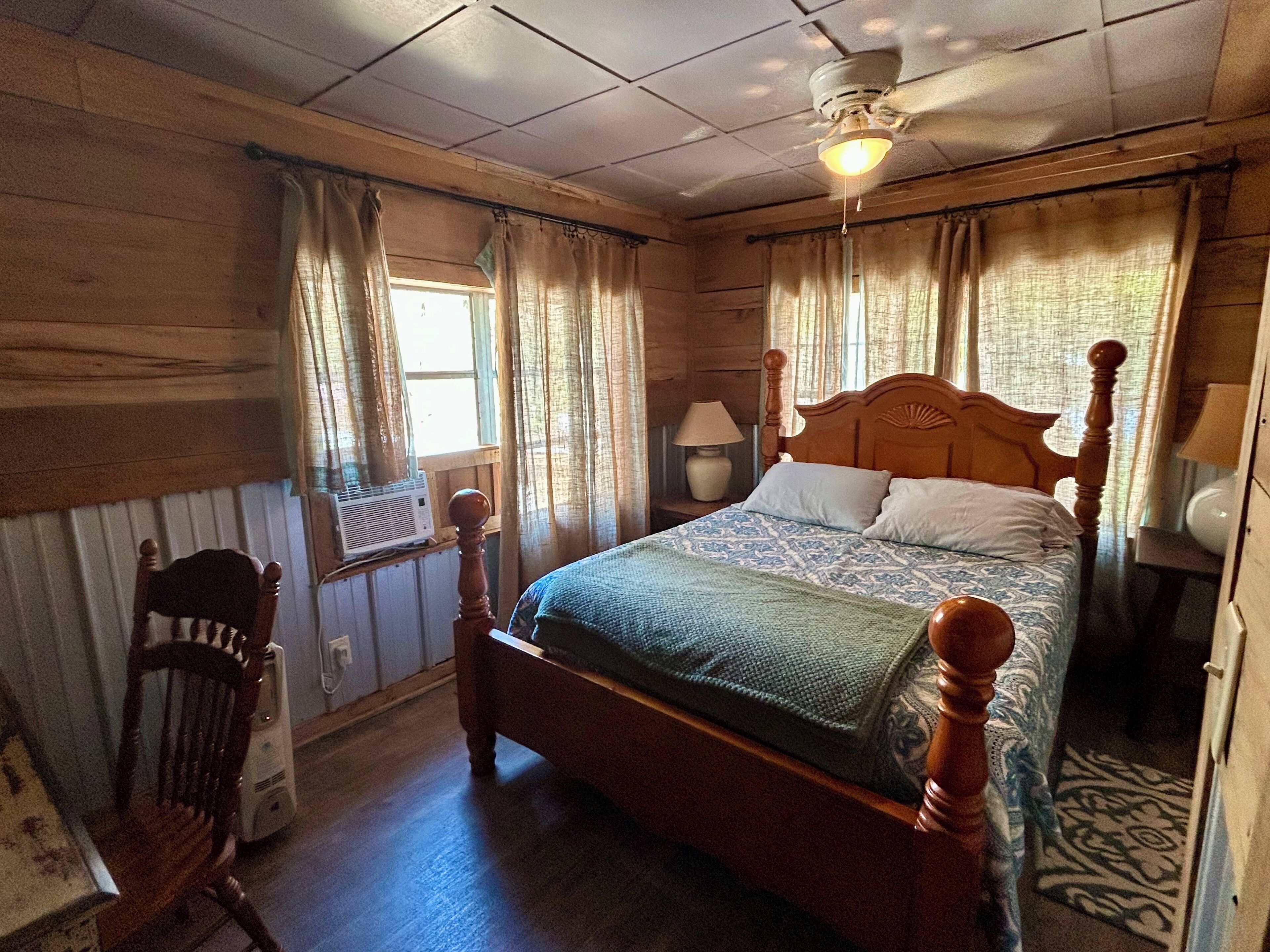 A wooden-paneled room with a single bed, a chair, and a ceiling fan.