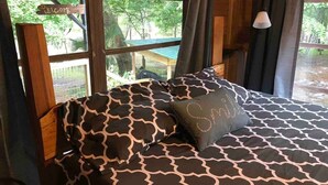 A bed with a patterned bedspread and a pillow with the word "Smile" on it, set in a room with wooden walls and a window overlooking greenery. - Bowman's Landing Spring House on the Santa Fe River (Fort White)