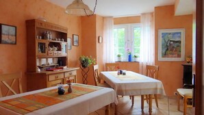 Un coin repas avec deux tables, un buffet en bois contenant de l’ustensile de cuisine et une fenêtre avec des rideaux.