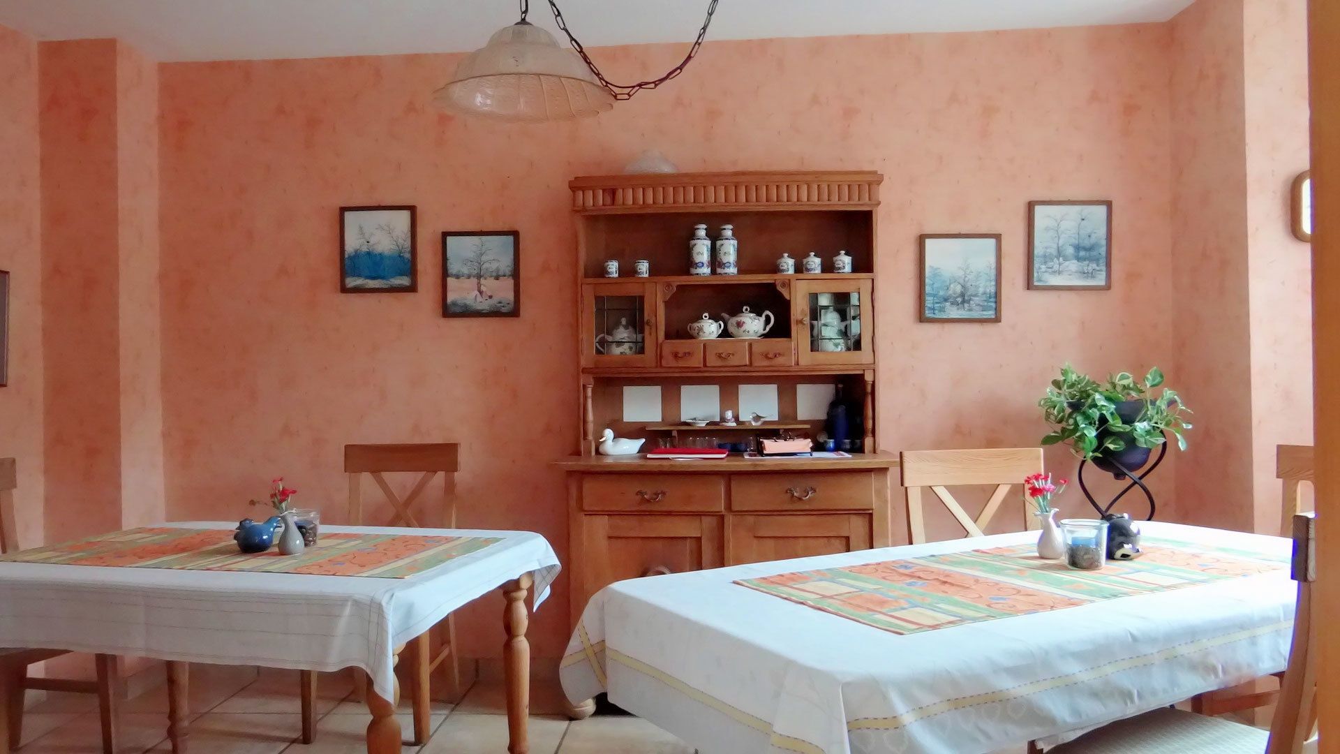 A dining area with two tables, chairs, a wooden cabinet with kitchenware, and framed pictures on the wall.