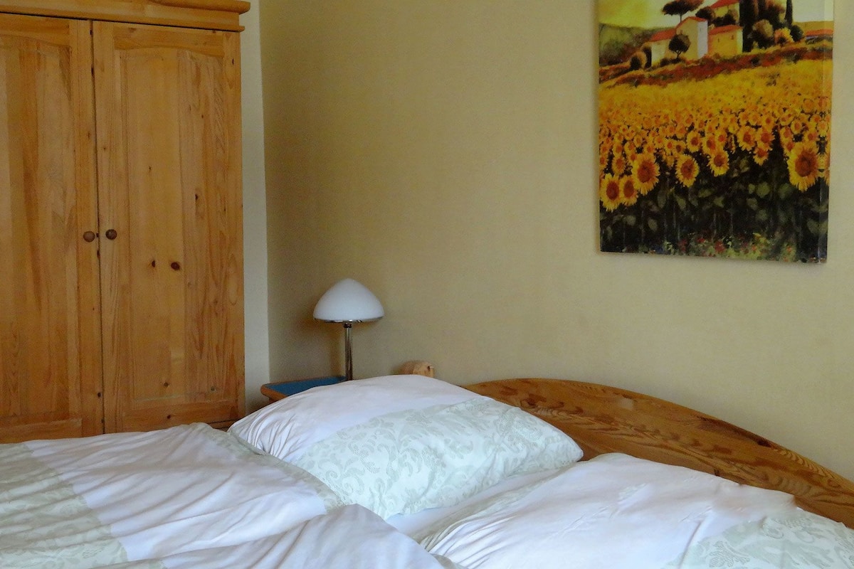A bedroom with a wooden wardrobe, a bed with white bedding, and a painting of sunflowers on the wall.