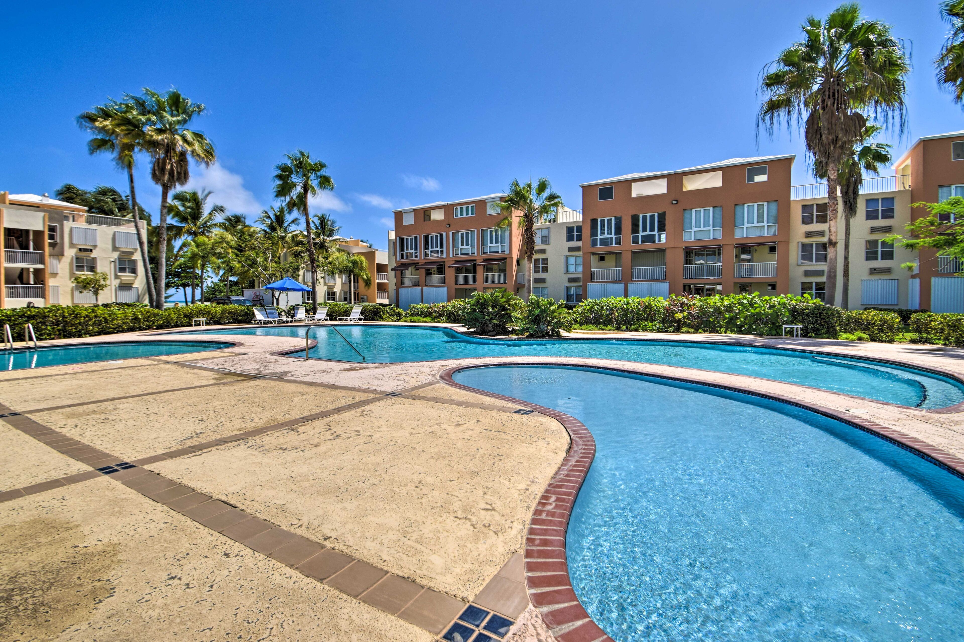 A swimming pool with a sandy area, surrounded by palm trees and multi-story buildings with balconies.