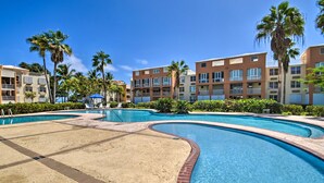 A swimming pool with a sandy area, surrounded by palm trees and multi-story buildings with balconies. - Loiza Condo w/ Rooftop Patio - Walk to Beach! (Loiza)