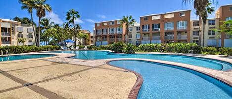 A swimming pool with a sandy area, surrounded by palm trees and multi-story buildings with balconies.