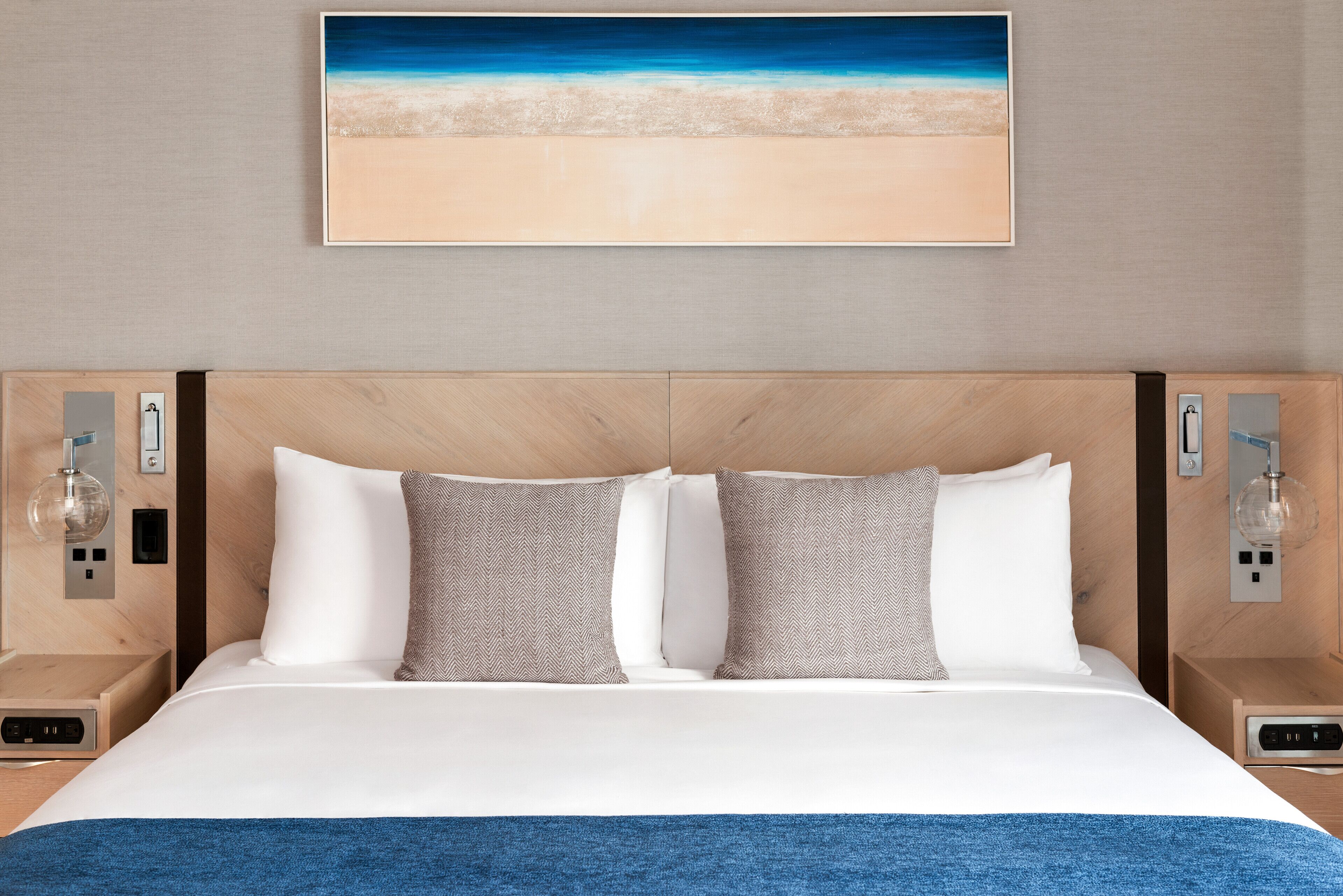 Una cama bien hecha con almohadas blancas y estampadas, una manta azul y un cuadro con motivos playeros.
