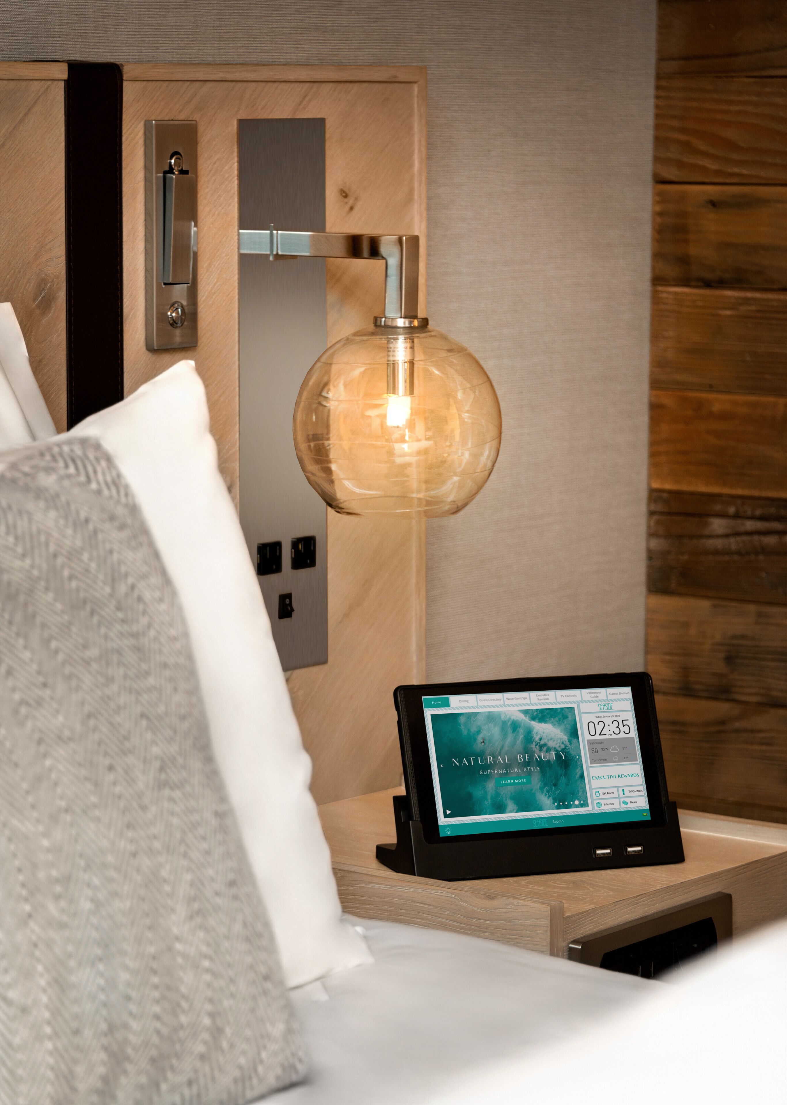 Habitación de hotel con cama, mesita de noche con una tablet que muestra un sitio web, lámpara de pared y panel de pared de madera.