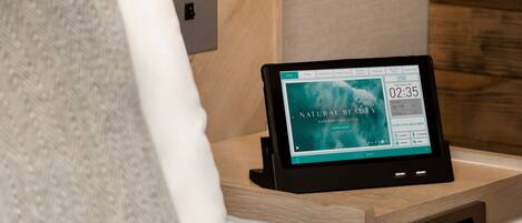 Habitación de hotel con cama, mesita de noche con una tablet que muestra un sitio web, lámpara de pared y panel de pared de madera.