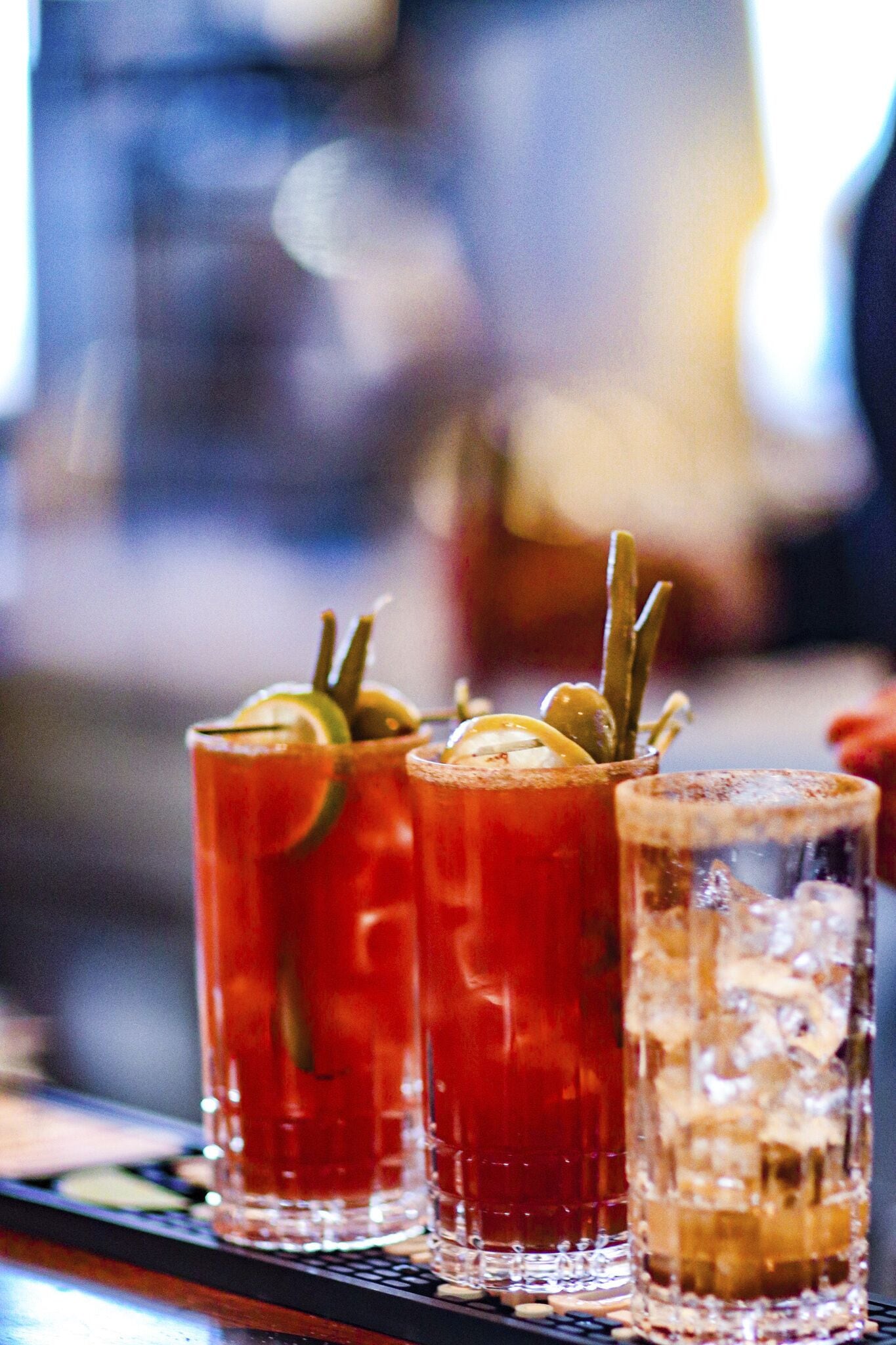Three glasses of a red beverage, possibly a Bloody Mary, with garnishes, on a bar counter.