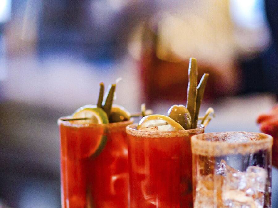 Three glasses of a red beverage, possibly a Bloody Mary, with garnishes, on a bar counter.