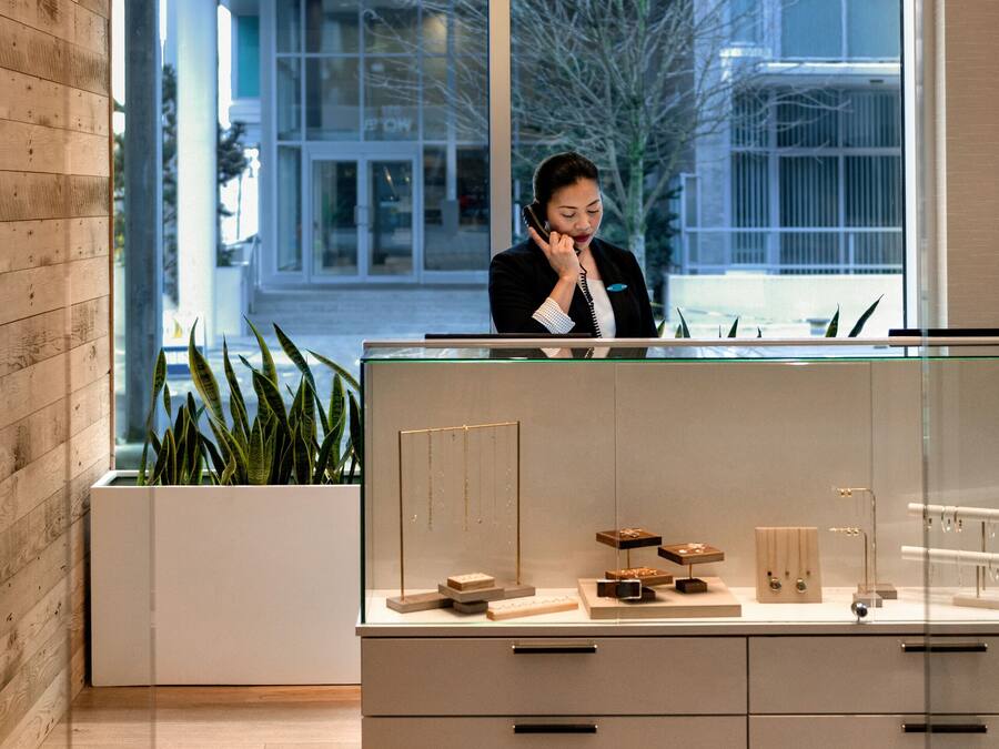 Ein Empfangsbereich mit Rezeptionistin, einem großen Fenster mit Blick auf ein Gebäude und einer Pflanze in der Ecke.