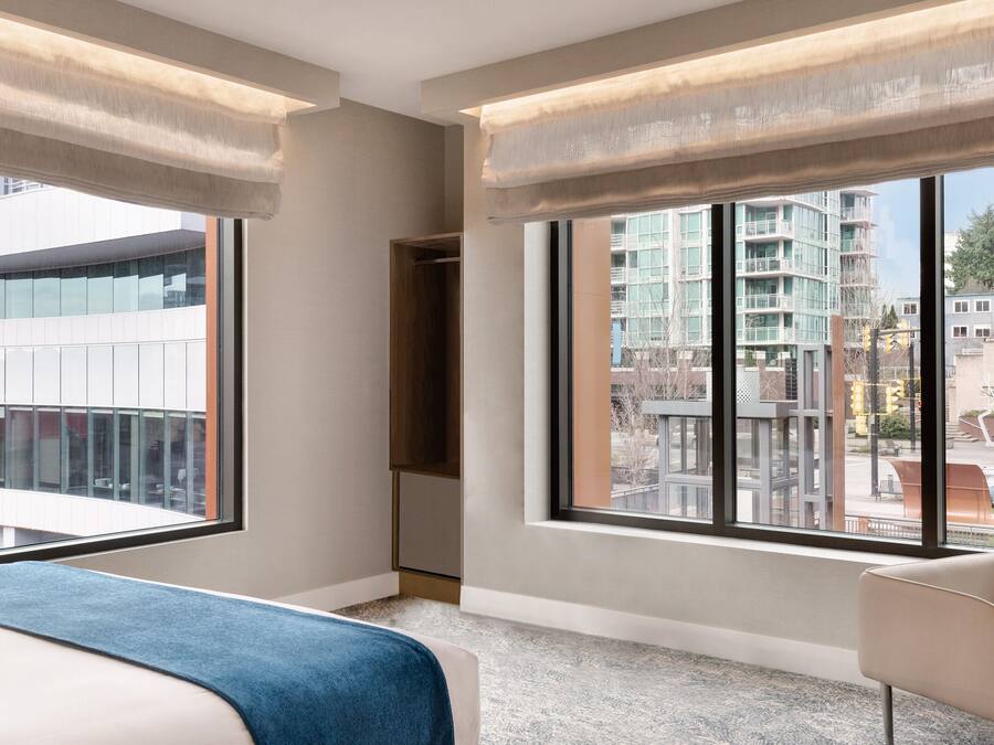 A modern hotel room with large windows, a bed, a desk, and a chair.