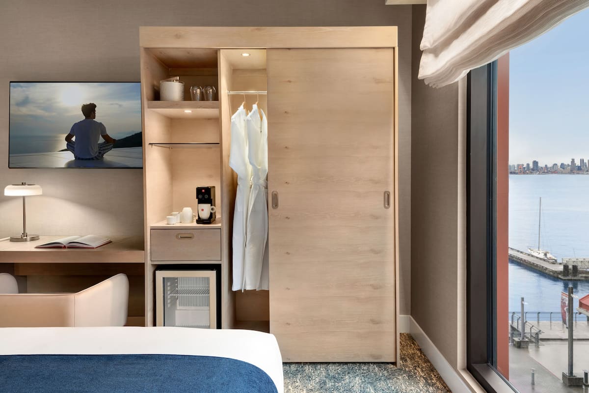 Hotellihuone, jossa on sänky, työpöytä kannettavalla tietokoneella, televisio, vaatekaappi sekä näkymä kaupunkiin ja vesistöön.