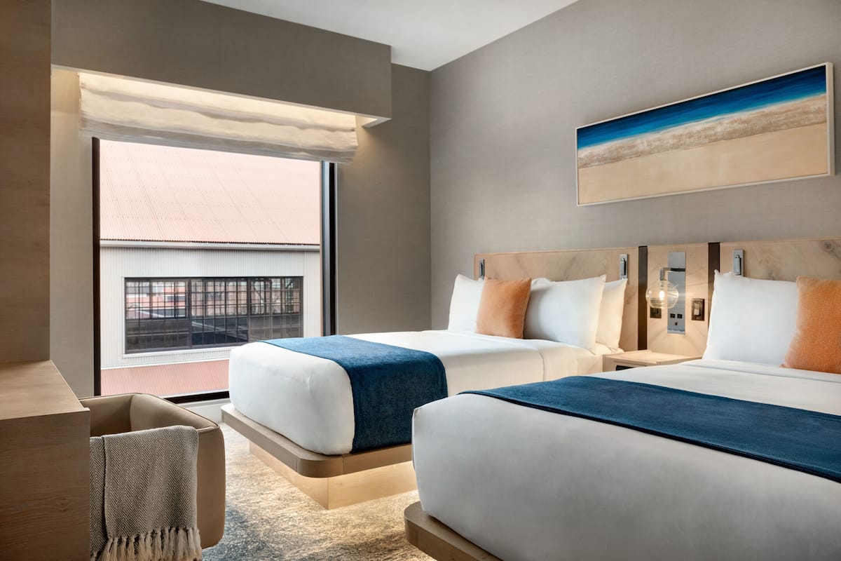 Moderni hotellihuone, jossa on kaksi sänkyä, suuri ikkuna, josta on näkymä rakennukseen, ja taulu seinällä.