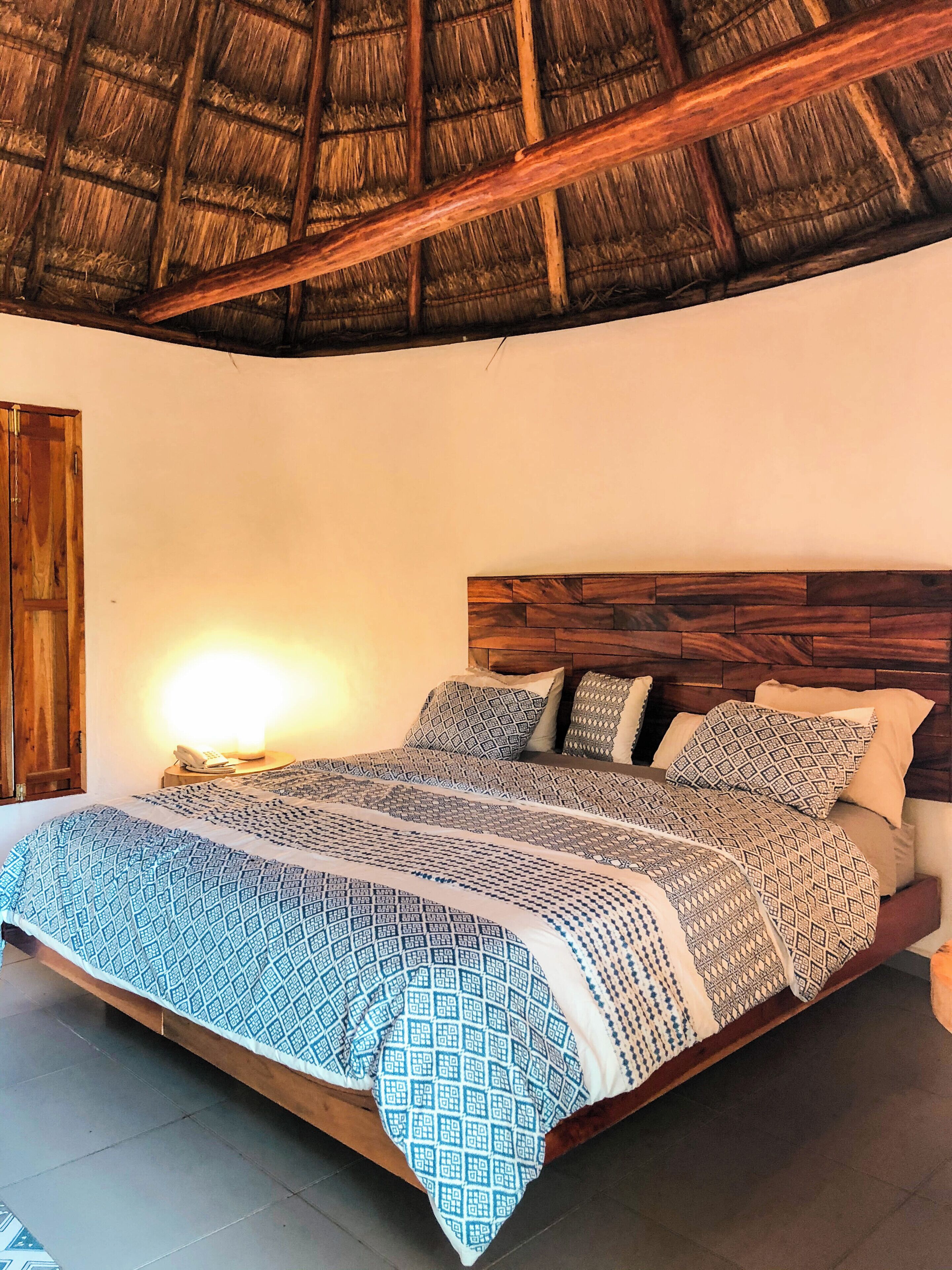 A bedroom with a wooden headboard, a patterned bedspread, and a thatched roof ceiling.