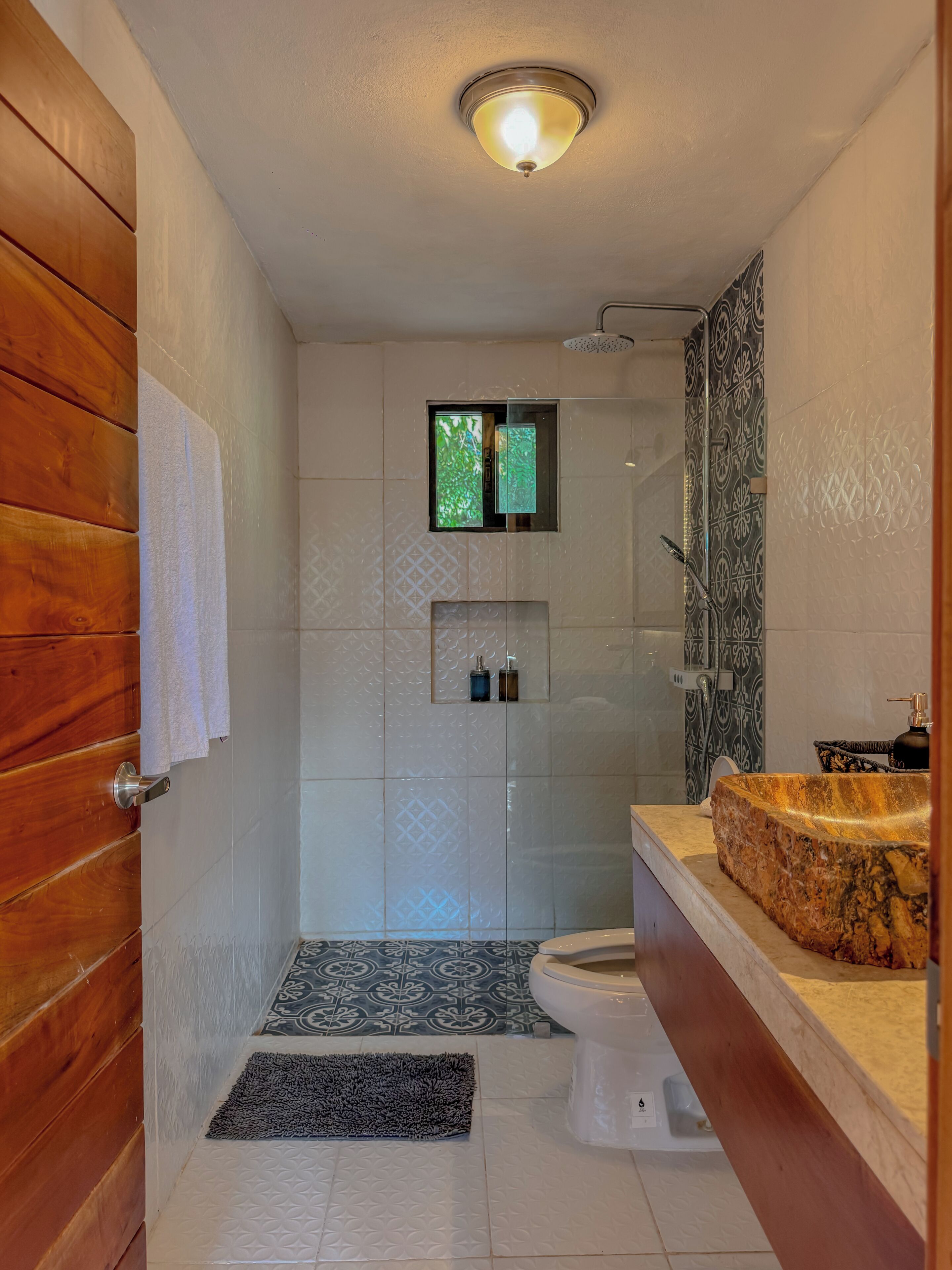 A bathroom with a wooden door, a white toilet, a shower with a glass door, a sink with a granite countertop, and patterned tiles.