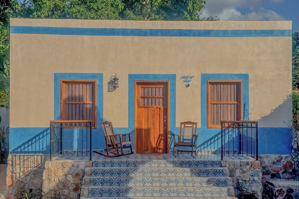 A building with a blue and beige facade, a wooden door, two windows, and a set of stairs leading to the entrance.