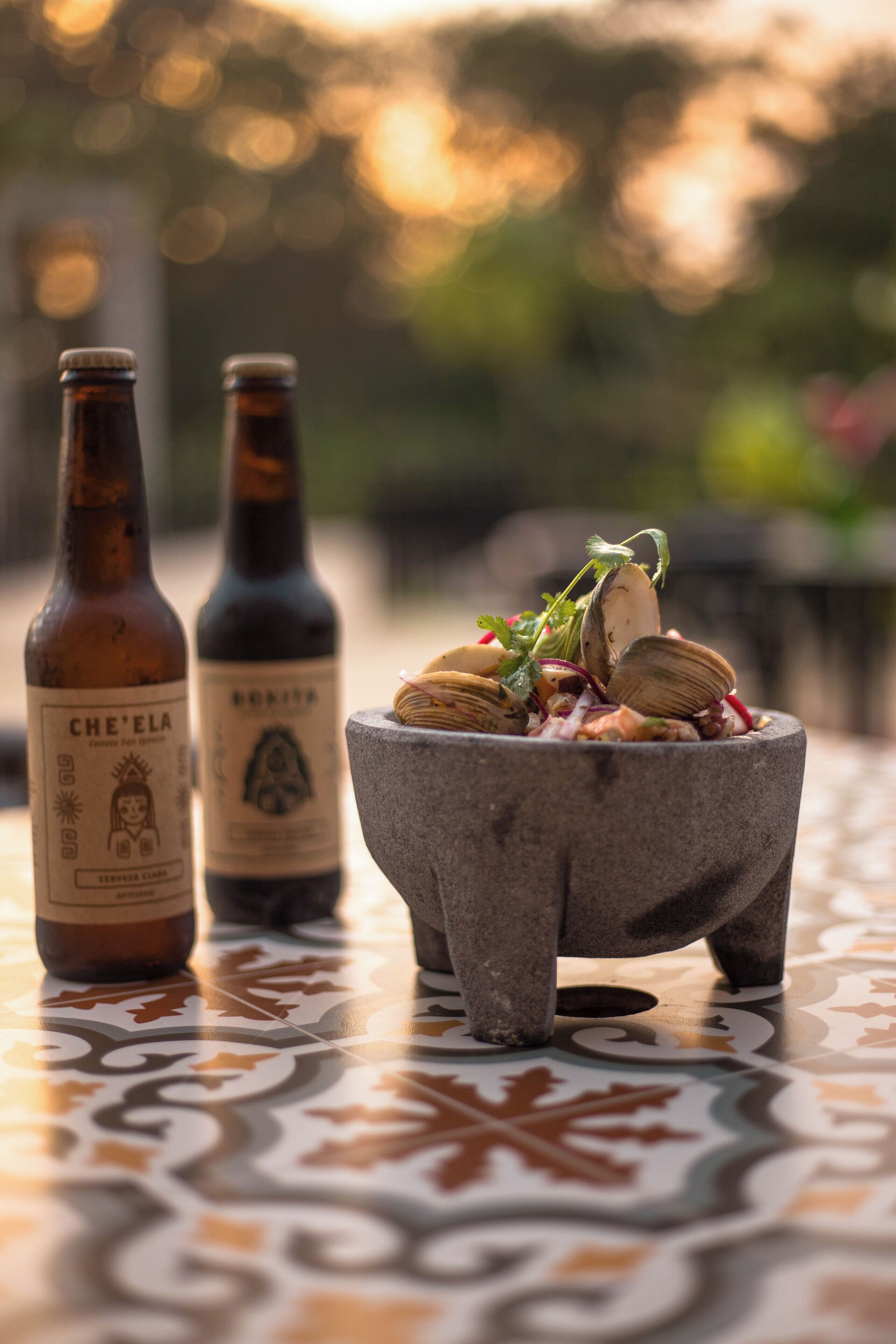 Two bottles of beer and a stone bowl with clams on a tiled table.