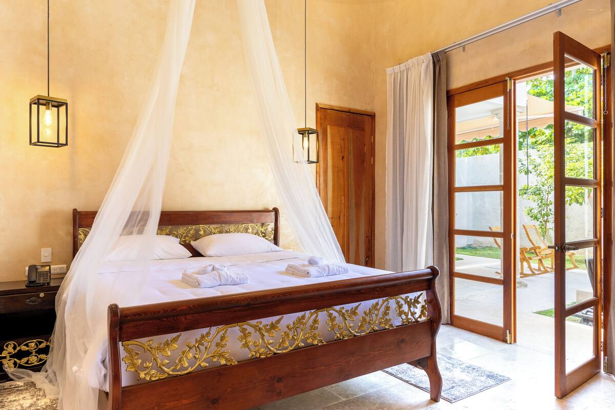 A bedroom with a wooden bed, white bedding, a canopy, and a view through an open door.