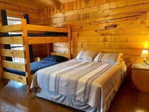Habitación tipo cabaña de madera con literas y una cama individual, suelo de madera y una mesita de noche con lámpara.