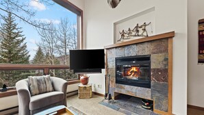 Ein modernes Wohnzimmer mit Kamin aus Stein, einer gemütlichen Couch, einem Flachbildfernseher und Blick auf Bäume durch große Fenster.