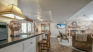 A modern kitchen with a bar area, wooden chairs, a fireplace, and a stone wall. - Riverside Home w/Private Hot Tub, Fire Pit & Deck! (Keystone)