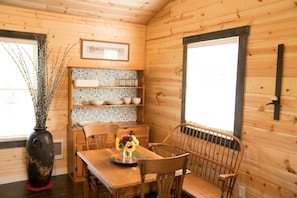 Interior de una cabaña de madera con una mesa de comedor, sillas, un jarrón con ramas, un revoque de azulejos y una ventana.
