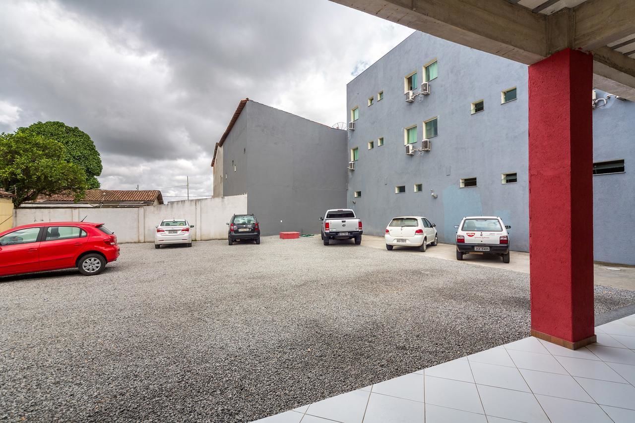 A parking area with several cars, a building with a grey facade and red pillar, and a tree in the background.