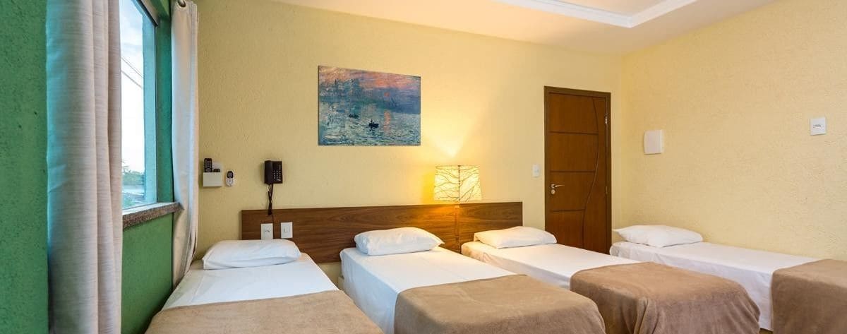 Een hotelkamer met drie eenpersoonsbedden, een houten hoofdbord, een schilderij aan de muur, een bedlampje en een raam met gordijnen.