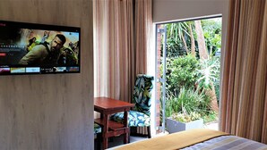 A hotel room with a large TV on the wall displaying a movie, a bed, a small table, a chair, and a view of a garden through a sliding door. - Hadeda Hall (Johannesburg)