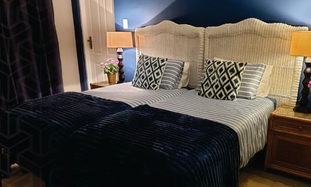 A bed with a striped bedspread and two pillows with geometric patterns, a woven headboard, a bedside table with a lamp, and a wall with a blue accent.