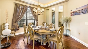 A dining room with a round table, ornate chairs, a chandelier, and a large window with curtains. - Stunning Large 7 bed Home With Private Pool, Spa, Balcony, Cinema Room, Fitness Suite and Able to see the Disney Fireworks - 467 (Kissimmee)
