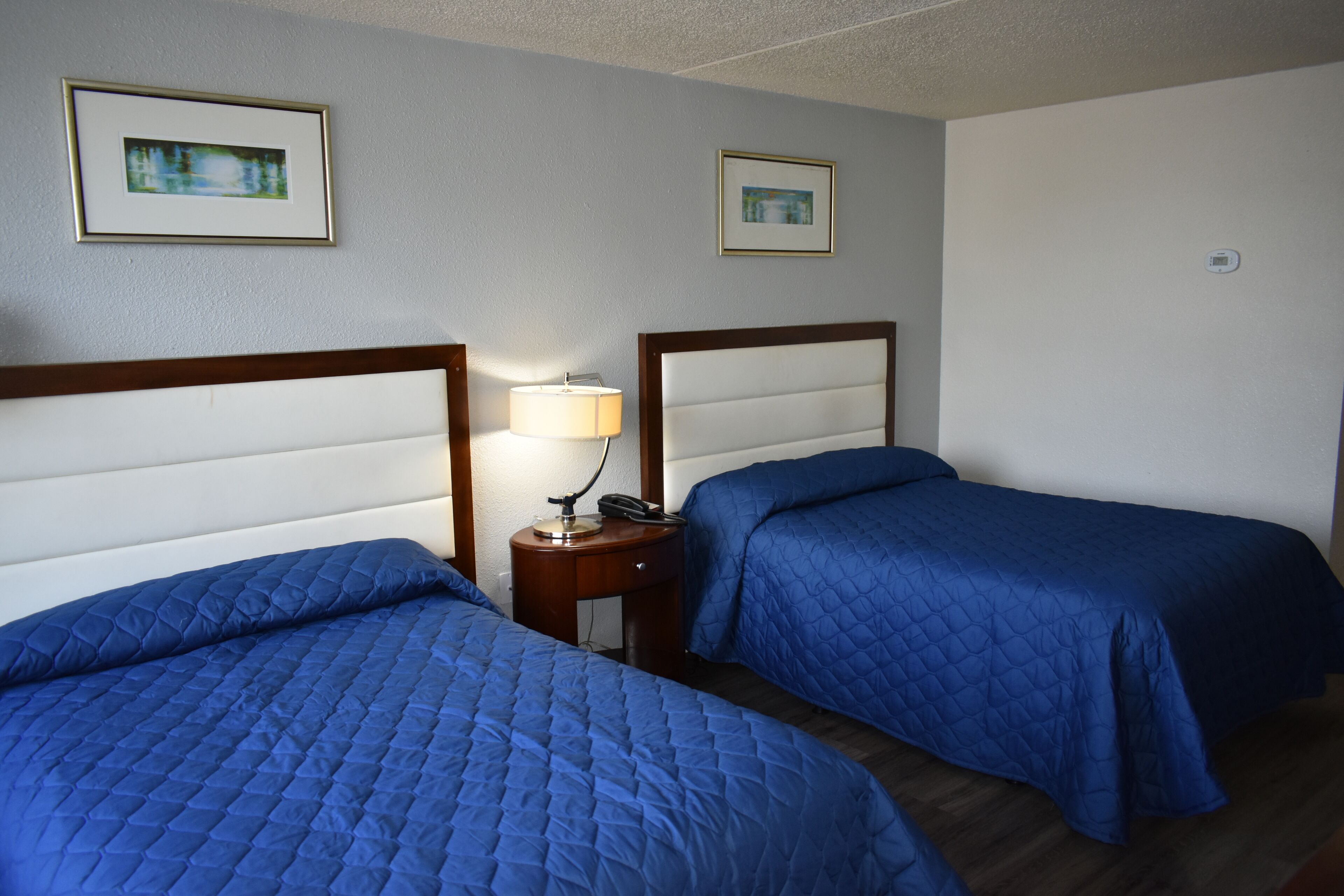 Una camera d'albergo con due letti, ciascuno con copripiumino azzurro trapuntato, un comodino con una lampada e quadri appesi alle pareti.