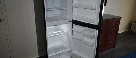 A compact refrigerator with multiple shelves and drawers, situated next to a wooden dresser and a bed in a room with a wooden floor and a white wall.